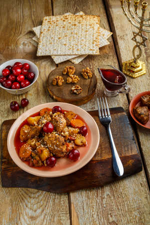 Jewish dish stew with chicken in cherry sauce decorated with cherries on a stand next to matzo, fork and ingredients.の写真素材