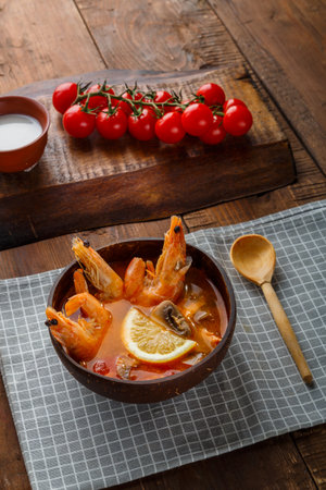 Tom yam soup with shrimps and coconut milk on a table on a checkered napkin next to vegetables and a wooden spoon.の写真素材