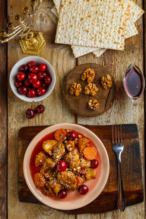 Jewish dish stewed potatoes with chicken in cherry sauce decorated with cherries on the table in a plate next to matzo and menorah. Vertical photoの写真素材