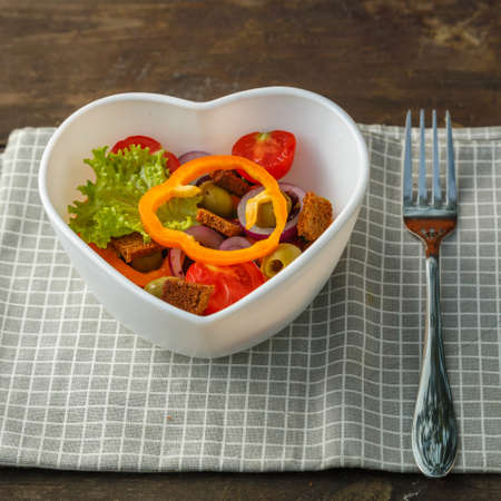 Vegetable salad in a heart shape plate on a wooden table on a checkered napkin next to a fork. Square photoの写真素材