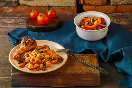 Seashell pasta in a tomato with a chicken leg baked on a grill on a blue napkin next to a fork and a vegetable salad. Horizontal photoの写真素材