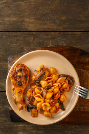 Seashell pasta in tomato and grilled chicken leg on a stand. Vertical photoの写真素材