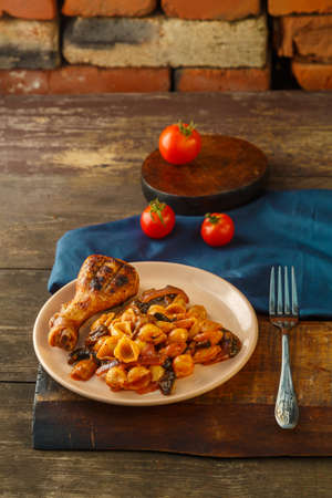 Seashell pasta in a tomato with a chicken leg baked on a grill on a stand and a napkin next to tomatoes and a vegetable salad. Vertical photoの写真素材
