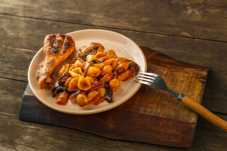 Seashell pasta in tomato and grilled chicken leg on a wooden stand next to a fork. Horizontal photoの写真素材