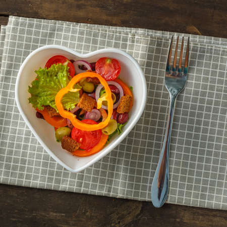 Vegetable salad in a heart shape plate on a wooden table on a checkered napkin next to a fork.の写真素材