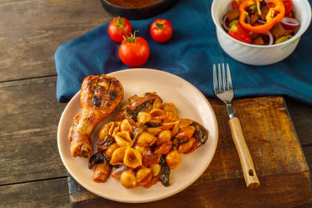 Seashell pasta in a tomato with a chicken leg baked on a grill on a blue napkin next to a fork and a vegetable salad.の写真素材