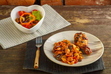 Seashell pasta in a tomato with a grilled chicken leg on a stand and a napkin next to a fork and a vegetable salad.の写真素材