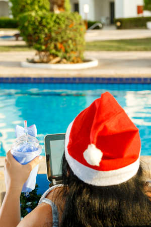 A woman in a Santa hat congratulates online friends with a laptop in a sun lounger by the pool, back view. Vertical photoの写真素材