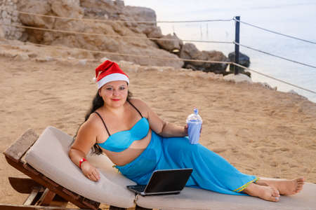 A woman on the seashore in a sun lounger in a santa claus hat drinks a cocktail and wishes a happy new year from a laptop.の写真素材