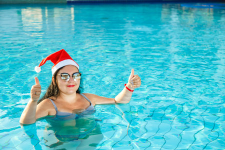 A woman in a Santa hat in the pool celebrates Christmas and shows her class with her hand.の写真素材