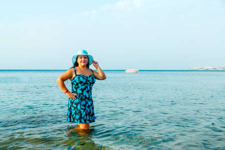 A happy Jewish woman in a blue sundress and hat stands in the sea knee-deep in water. Horizontal photoの写真素材