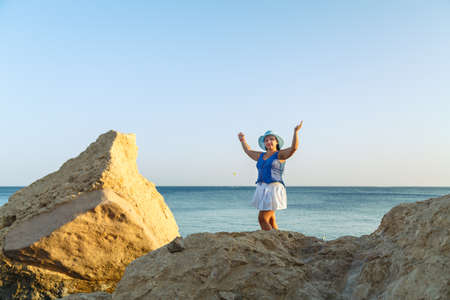 A young brunette woman in a white skirt and a sun hat on the seashore stands against the background of the sea and sky. Horizontal photo.の写真素材