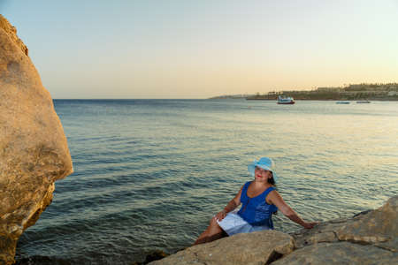 A young Jewish woman in a white skirt and a sun hat on the seashore sits on a rock. Horizontal photo.の写真素材