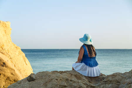 A young brunette woman in a white skirt and blue hat on the seashore sits on the rocks with her back to the camera.の写真素材