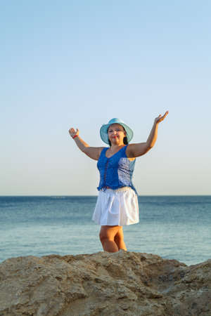 Young Jewish woman in a white skirt and blue hat on the seashore near the rocks happy.の写真素材