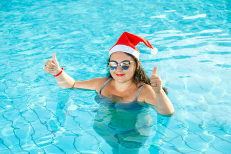 A woman in a Santa hat in the pool celebrates Christmas and shows her class with her hand. Horizontal photoの写真素材