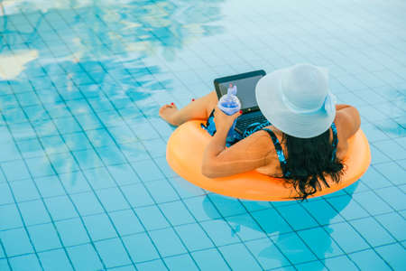 Brunette woman in a sun hat in the pool in a swimming circle with a cocktail and a laptop with her...
