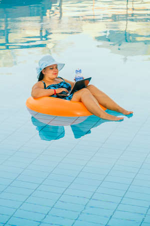 A woman in a sun hat in the pool in a swimming circle with a cocktail and a laptop communicates via...