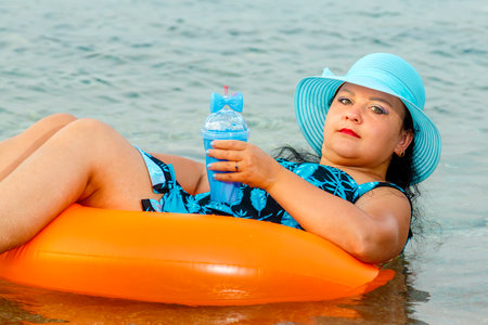 A woman in a straw hat and glasses in the sea in a swimming circle with a cocktail in her hand. Horizontal photo.の写真素材