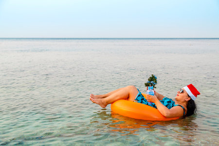 A woman in a swimming ring in a Santa Claus hat with a cocktail and a small Christmas tree in her hand floats in the sea during Christmas holidays. Horizontal photo.の写真素材