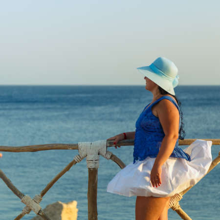 A young woman in a white skirt and a sun hat stands on the shore against the backdrop of the sea at sunset looks at the sea. Square photoの写真素材