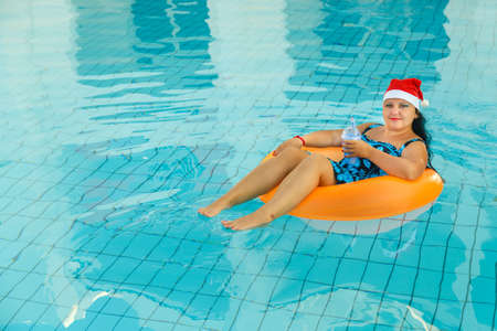 Young brunette woman in santa claus cap in a swimming circle in the pool with a cocktail in hand in the new year holidays. Horizontal photoの写真素材