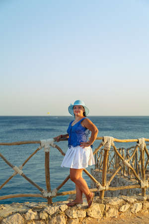 Young brunette woman in a white skirt and blue hat on the seashore happy. Vertical photoの写真素材