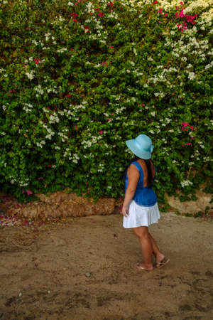 A brunette woman in a white skirt and a sun hat stands by the blooming white bougainvillea with her back to the camera. Vertical photoの写真素材