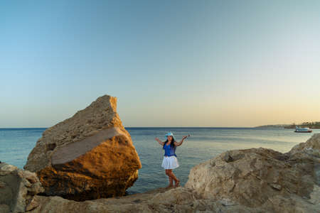 A young woman in a white skirt and a sun hat stands on the shore among the rocks against the backdrop of the sea at sunset. Horizontal photoの写真素材