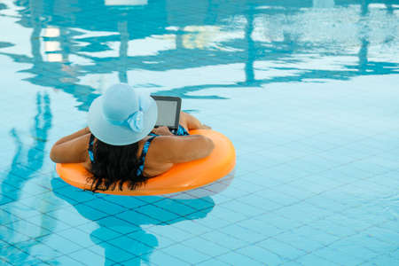 Woman in a sun hat in a swimming pool in a swimming circle with a laptop relaxes shooting from the back. Horizontal photoの写真素材
