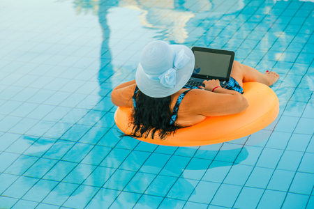 Brunette woman in a sun hat in the pool in a swimming circle with a cocktail and a laptop with her back to the camera. Horizontal photoの写真素材