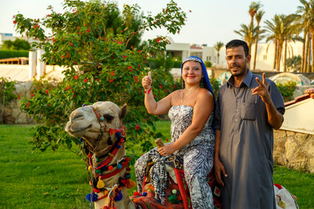 Egypt, Sharm El Sheikh, August 30, 2021 a woman in a cape on her head next to a Bedouin and a camel and lying on the grass on a city street.のeditorial素材