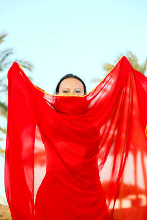 A woman, an oriental dancer, against a background of palm trees with a closed cape, her face, only her eyes are visible.の写真素材