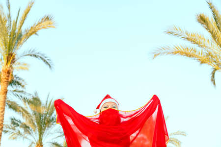A woman-dancer of oriental dance in a Santa hat on a background of palm trees with a closed cape over her face.の写真素材