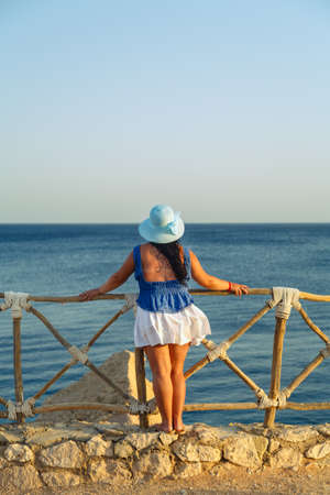 A young brunette woman in a white skirt and a blue hat on the seashore admires the sea with her back to the camera. Vertical photoの写真素材