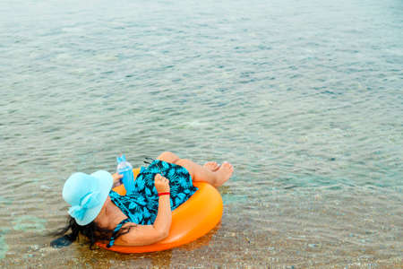 Side view of a woman in a straw hat and glasses in the sea in a swimming circle with a cocktail in...