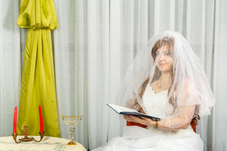 The Jewish bride sits at the table before the Hupa ceremony in a wedding dress, covering her face...