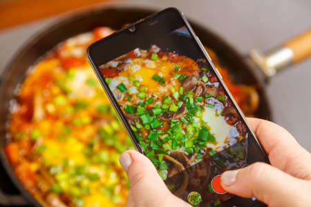 The phone in female hands takes pictures of shakshuka in a frying pan. horizontal photoの写真素材