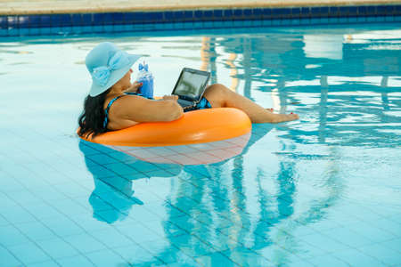 Woman in a sun hat in a swimming pool in a swimming circle with a laptop relaxes filming from the side. horizontal photoの写真素材