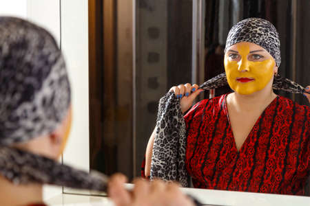 A bald Jewish woman on her face with a cosmetic mask puts on a traditional headdress in front of a...