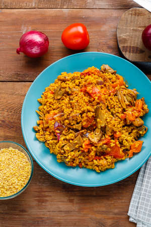 A large dish of bulgur with vegetables and mushrooms in spices on a blue napkin next to tomatoes onions on the table. vertical photoの写真素材