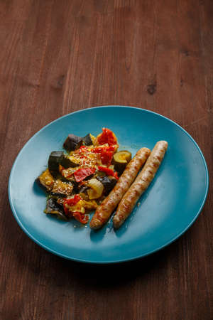 A plate of vegetables tomatoes zucchini eggplant with grilled sausages on a wooden table. vertical photoの写真素材