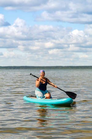 a woman in a closed swimsuit, a pareo and a headdress on her knees on a SUP board with a paddle...