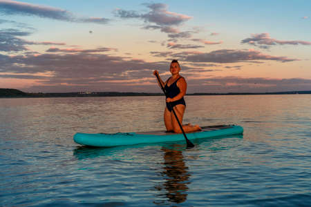 a woman in a closed swimsuit on her knees on a SUP board with an oar floats on the water against the...