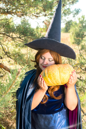 A red-haired witch in a pointed hat and cloak in the forest with a candle in an old candlestick and a pumpkin in her hands. vertical photoの写真素材
