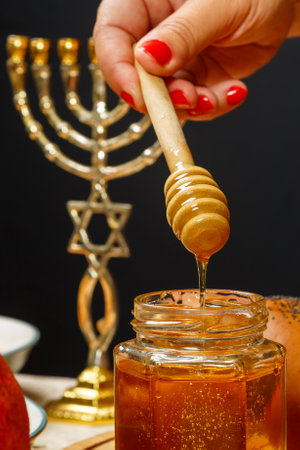 Honey drips from a wooden spoon into a jar next to the menorah challah and pomegranate. vertical photoの写真素材