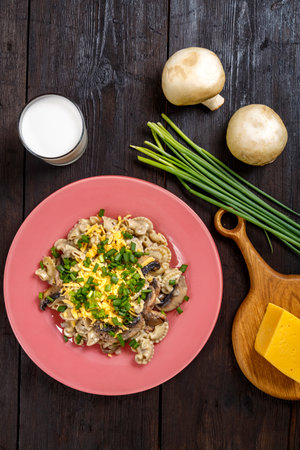 Pasta in a creamy sauce with mushrooms in a plate on the table next to a glass of cream, cheese on a wooden board, green onions and champignons top view. vertical photoの写真素材