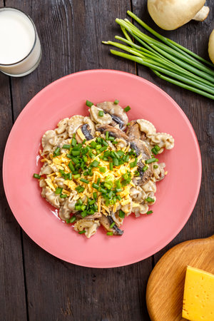 Pasta in a creamy sauce with mushrooms in a plate on the table next to a glass of cream, green onions and champignons. vertical photoの写真素材