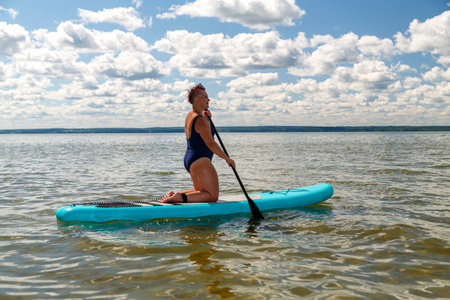 a woman in a closed swimsuit with a mohawk on her knees on a SUP board with an oar floats on the water. horizontal photoの写真素材