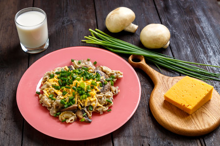 Pasta in a creamy sauce with mushrooms in a plate on a dark wooden table next to a glass of cream, cheese on the board, green onions and mushrooms. horizontal photoの写真素材